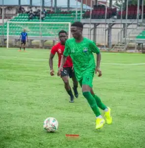 Soccer player in a green kit with number 14 dribbles the ball on a grassy field, pursued by a defender in red; stadium stands in the background.