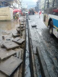 Urban street scene showing a broken sidewalk with open trench and scattered concrete slabs along a flooded, muddy road; pedestrians nearby and a bus on the right.