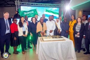 Diverse group of professionals posing around a large cake on a white-clothed table at a formal celebration, with Saudi and Nigerian flags in the background.