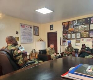 Group of soldiers in camouflage seated around a conference table in a briefing room with framed photos and posters on the walls.