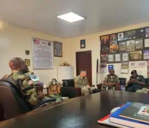 Group of soldiers in camouflage seated around a conference table in a briefing room with framed photos and posters on the walls.