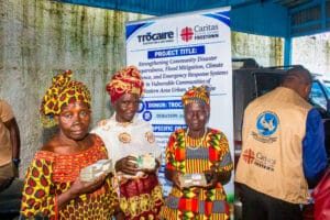 Three women in vibrant traditional outfits hold stacks of cash at a charity event, with a Trocaire–Caritas banner behind them.