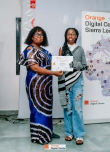 Two women exchange a certificate at a ceremony, standing beside an Orange Digital Center banner. (certificate reads 'Certificate of Completion')