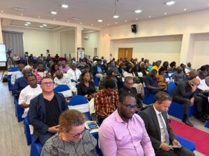 A crowded conference hall with attendees seated in blue chairs, listening to a presentation at the front of the room.