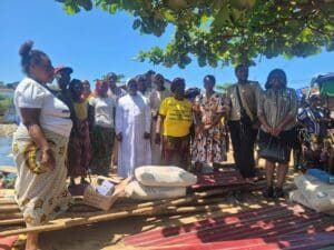 Group of men and women standing together under a leafy tree on a sunny day, with construction materials (cement bags and wooden planks) in the foreground behind them.