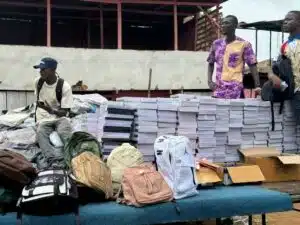Group of young men with backpacks around a display of stacked books and boxes outdoors, suggesting a book distribution or market setting.
