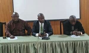 Three men seated at a conference table in a meeting room, the center man in a suit with a green tie holding a phone.