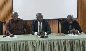 Three men seated at a conference table in a meeting room, the center man in a suit with a green tie holding a phone.