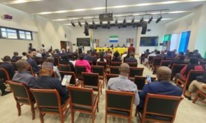 Audience seated in a conference hall facing a panel at a long yellow table on stage.