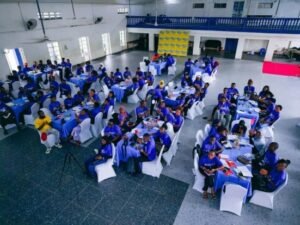 Large hall filled with attendees wearing blue shirts seated at round tables with white chairs in a banquet-style event.