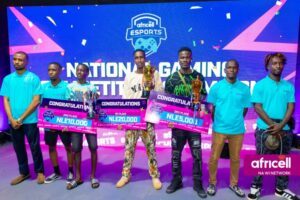 Six young men stand on a stage holding oversized prize checks and trophies at an Africell Esports awards ceremony, with confetti and a backdrop reading 'National Gaming Competition.'