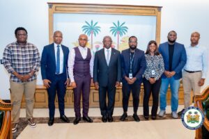 Eight professionals in suits and business attire pose for a formal group photo in a government building, with a palm-tree mural behind them and a seal in the corner