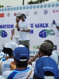 Speaker holding a microphone on a stage at a walk event, with a large banner and logos in the background.