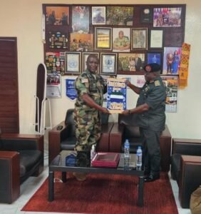 Two uniformed officers shake hands in a formal office, exchanging a blue and yellow certificate as wall photos and awards look on.