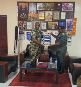 Two uniformed officers shake hands in a formal office, exchanging a blue and yellow certificate as wall photos and awards look on.