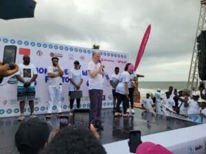 People on a stage by the coast, giving speeches to a crowd with banners and logos in the background under a cloudy sky.