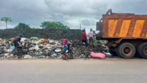 People sift through a roadside pile of garbage beside a large rusty dump truck on the right under a cloudy sky.