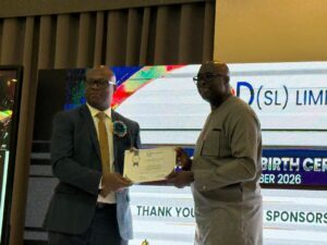 Two men on a stage exchange a certificate during an award ceremony; one wears a suit and yellow tie with a ribbon, the other in gray traditional attire.