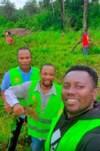 Three men in bright safety vests take a selfie in a lush tropical farming field with huts and palm trees in the background.