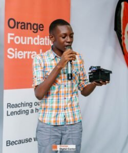 Young man in a plaid shirt speaks into a microphone while holding a small black device at an Orange Foundation Sierra Leone event.