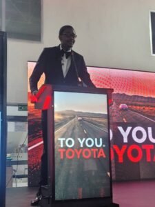 Man in a tuxedo delivering a talk at a podium, with a highway image and Toyota branding on the lectern screen behind him
