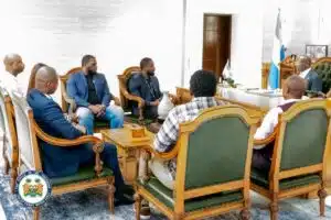 Six men in suits sit in ornate wooden chairs around a large desk in a presidential office; the presidential seal and flag are visible in the scene.