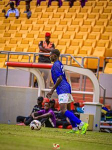 Soccer player in blue jersey (number 14) dribbles a ball on a grassy field near the sideline, with yellow stadium seats in the background.