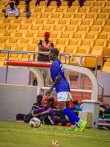 Soccer player in blue jersey (number 14) dribbles a ball on a grassy field near the sideline, with yellow stadium seats in the background.