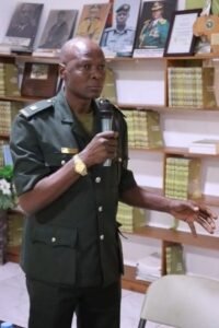 Uniformed officer speaks into a handheld microphone in a room lined with bookshelves and framed portraits in the background.