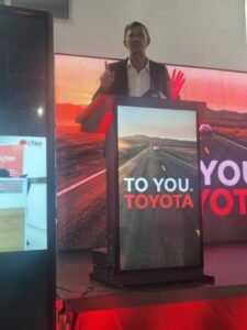 Male presenter in a suit speaks at a podium on a stage, microphone near mouth, with a red-lit backdrop reading 'TO YOU TOYOTA'.