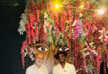Africell Backs Drizilik’s ‘Audacity’ as Sierra Leonean Music Targets Wider Audiences Two men posing in front of a vibrant flower-covered wall; one in a white suit with a burgundy hat and sunglasses, the other in a white shirt and light gray pants with black shoes.