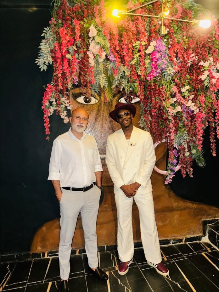 Two men posing in front of a vibrant flower-covered wall; one in a white suit with a burgundy hat and sunglasses, the other in a white shirt and light gray pants with black shoes.