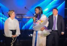 SLCAA DG Honoured at International Civil Aviation Organization Security Conference in Kazakhstan Woman in a traditional white dress holding a bouquet and microphone on stage, flanked by a woman in a white blouse and a man in a suit with blue stage lights behind.