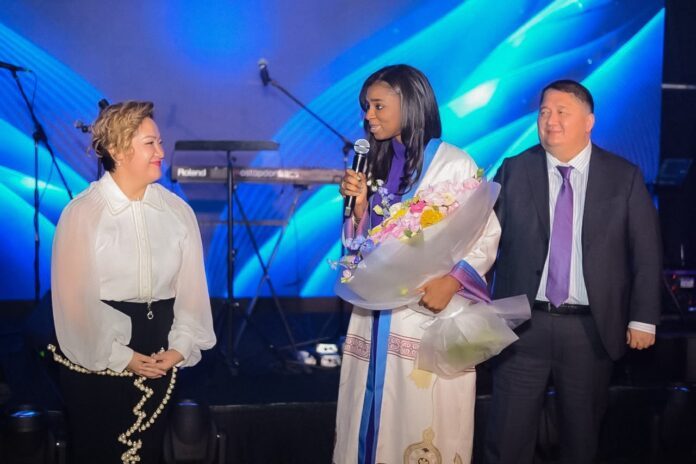 Woman in a traditional white dress holding a bouquet and microphone on stage, flanked by a woman in a white blouse and a man in a suit with blue stage lights behind.