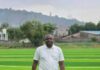 Ahead of 2026/2027 Season… Amb. Anthony Junior Navo Turns Vision into Reality as East End Lions Gets New Standard Pitch Man in a white polo shirt and dark jeans stands on bright green artificial turf outdoors, with trees and hills in the background.