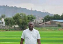 Ahead of 2026/2027 Season… Amb. Anthony Junior Navo Turns Vision into Reality as East End Lions Gets New Standard Pitch Man in a white polo shirt and dark jeans stands on bright green artificial turf outdoors, with trees and hills in the background.