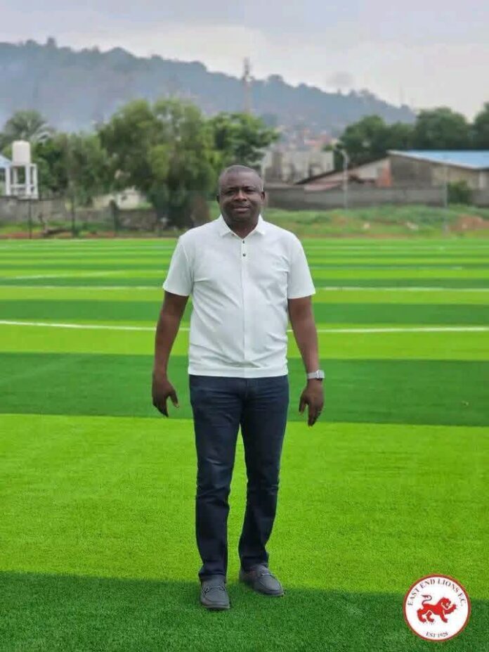 Man in a white polo shirt and dark jeans stands on bright green artificial turf outdoors, with trees and hills in the background.