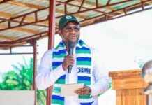Dr. Henry Musa Kpaka Commends Pee Cee & Sons for Transforming Sierra Leone’s Agricultural Landscape Man in white shirt and green cap speaks into a microphone on a wooden podium, wearing a blue-green striped scarf outdoors.”