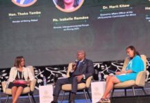 Mines Minister Pushes for Mineral Processing and Industrialization at Mining in Motion 2026 Three panelists seated on stage in beige chairs during a conference talk; the man in the center holds a microphone while two women listen, with bios projected on a large screen behind them.