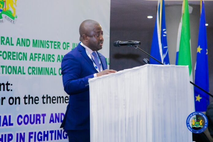 Male speaker in a blue suit delivering a talk at a white, draped podium with a microphone, backdrop shows a large green text banner and multiple flags nearby (official conference setting).