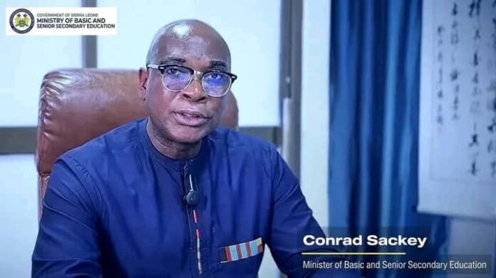 Conrad Sackey, Minister of Basic and Senior Secondary Education, seated in a blue shirt in an office, with the government of Sierra Leone logo in the top-left.