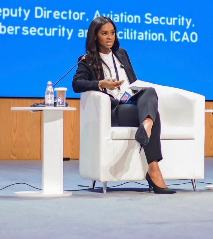 Woman panelist in a dark suit speaks into a microphone on stage, with a white chair, small table, and water bottle nearby.