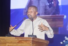 Vice President Urges Inclusive Governance, Greater Women’s Representation & PR to Consolidate Sierra Leone’s Peace Man speaking at a wooden podium on a stage with two microphones and a blue backdrop behind him, gesturing with his hands.