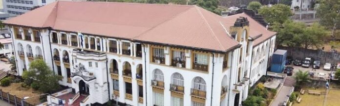 Aerial view of a large white colonial-style building with a red tile roof, arched windows and balconies, surrounded by trees and a parking area.