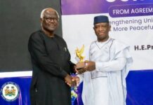 President Bio, Ernest Bai Koroma Unite in Call for National Peace, Reconciliation and Political Tolerance Two men shake hands on a stage as one presents a gold eagle trophy; a purple banner looms in the background.