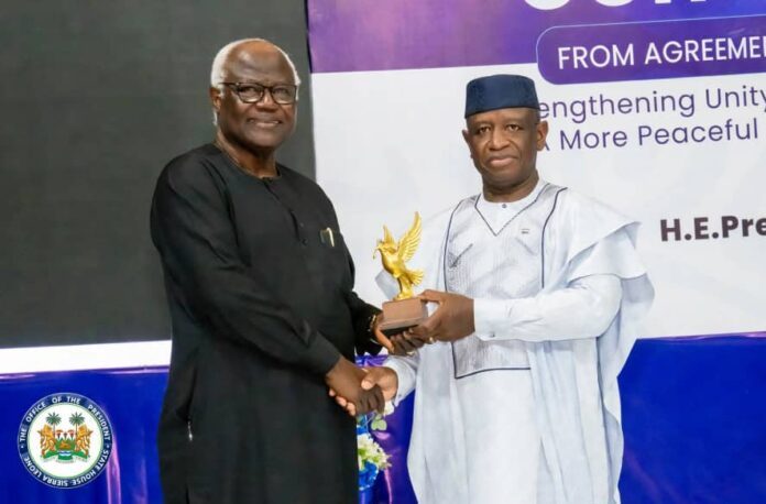 Two men shake hands on a stage as one presents a gold eagle trophy; a purple banner looms in the background.