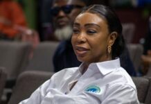 Hawa Sally Samai: The Woman Behind the National Conversations Shaping Sierra Leone’s Peace and Unity Woman with dark hair in a white button-down shirt, seated at an event, smiling slightly.