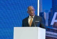 President Bio Urges African Leaders to Prioritize Economic Independence, Regional Trade and Accountable Governance African man in a suit speaks at a podium for the Elevate Africa event, with a blue backdrop behind him and city imagery on the side panel.