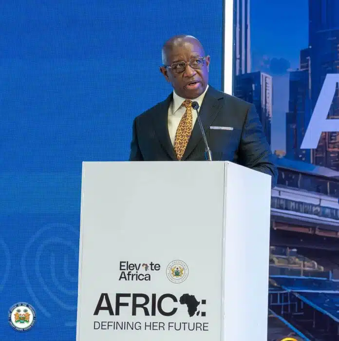 African man in a suit speaks at a podium for the Elevate Africa event, with a blue backdrop behind him and city imagery on the side panel.