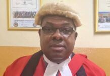 “I Am Not Joking!” Justice Adrian Fisher Warns Against Disruption of APC Internal Elections Portrait of a judge in a red judicial robe with white neckbands and a light wig, seated in an office with framed certificates on the wall and an official seal in the corner.